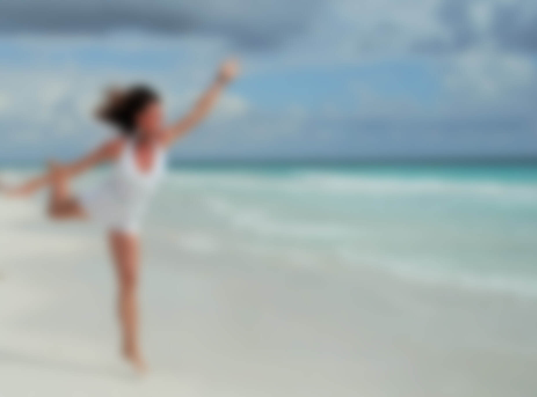 A woman in white with arms thrown up in a joyful balance on bright white sand beside turquoise shallows