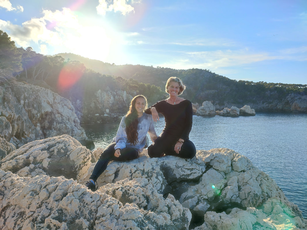Sandra Van Olmen with a younger woman, sitting on the rocks above a Mallorcan cove in warm late-afternoon light