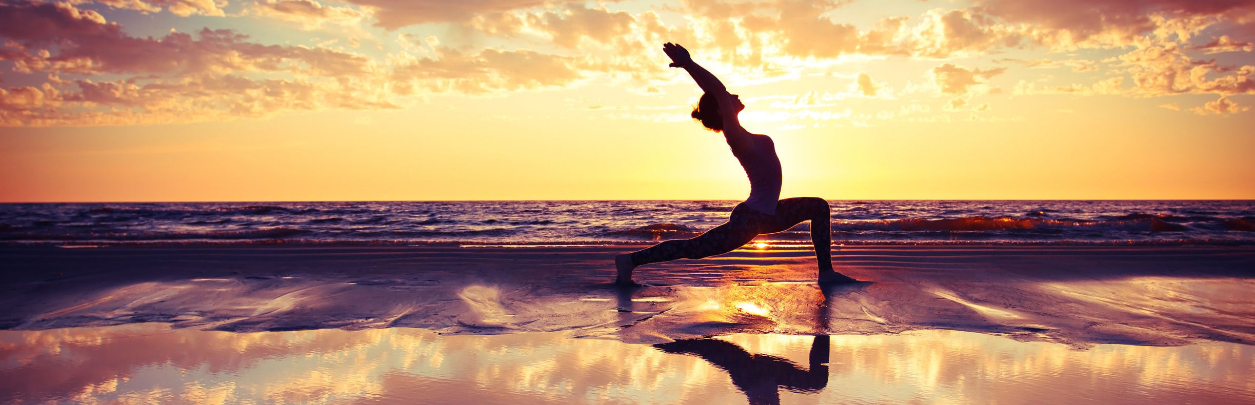 A single figure holds a yoga pose on wet sand at sunset, the warm sky and the sea mirrored in the water at their feet